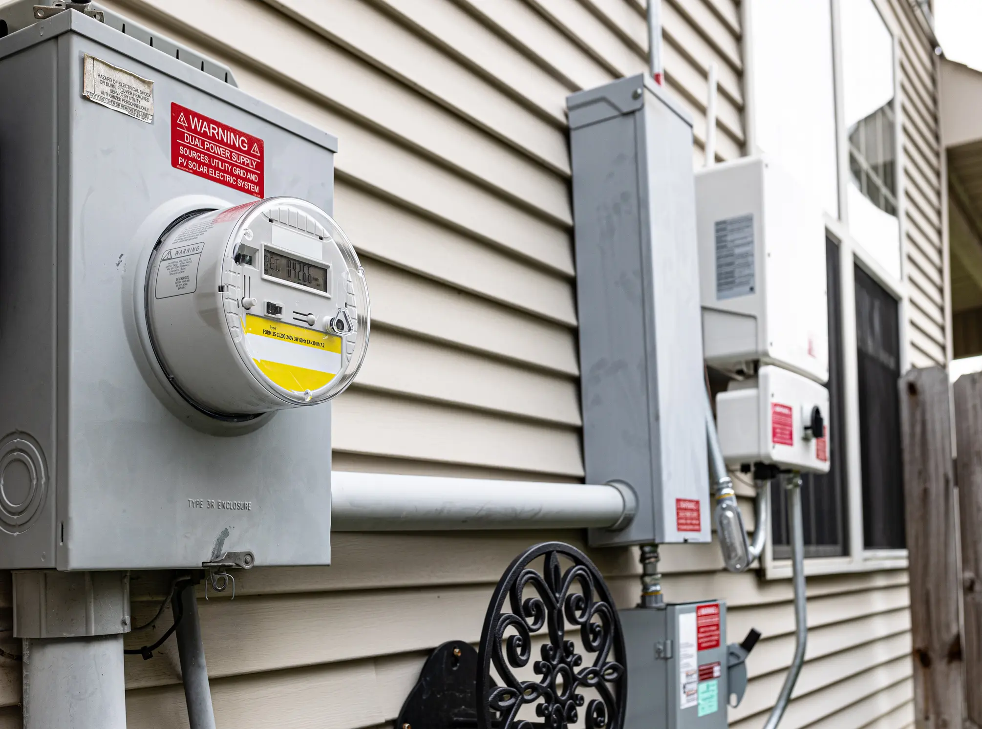 The utility electricity bill meter is connected to the photovoltaic system inverter and quick disconnect box, providing a dual power supply. Stickers indicating the solar panel source are placed according to regulations.